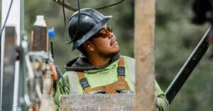 Construction worker in a hard hat and safety glasses looks up at a work site, holding a perforated metal panel.